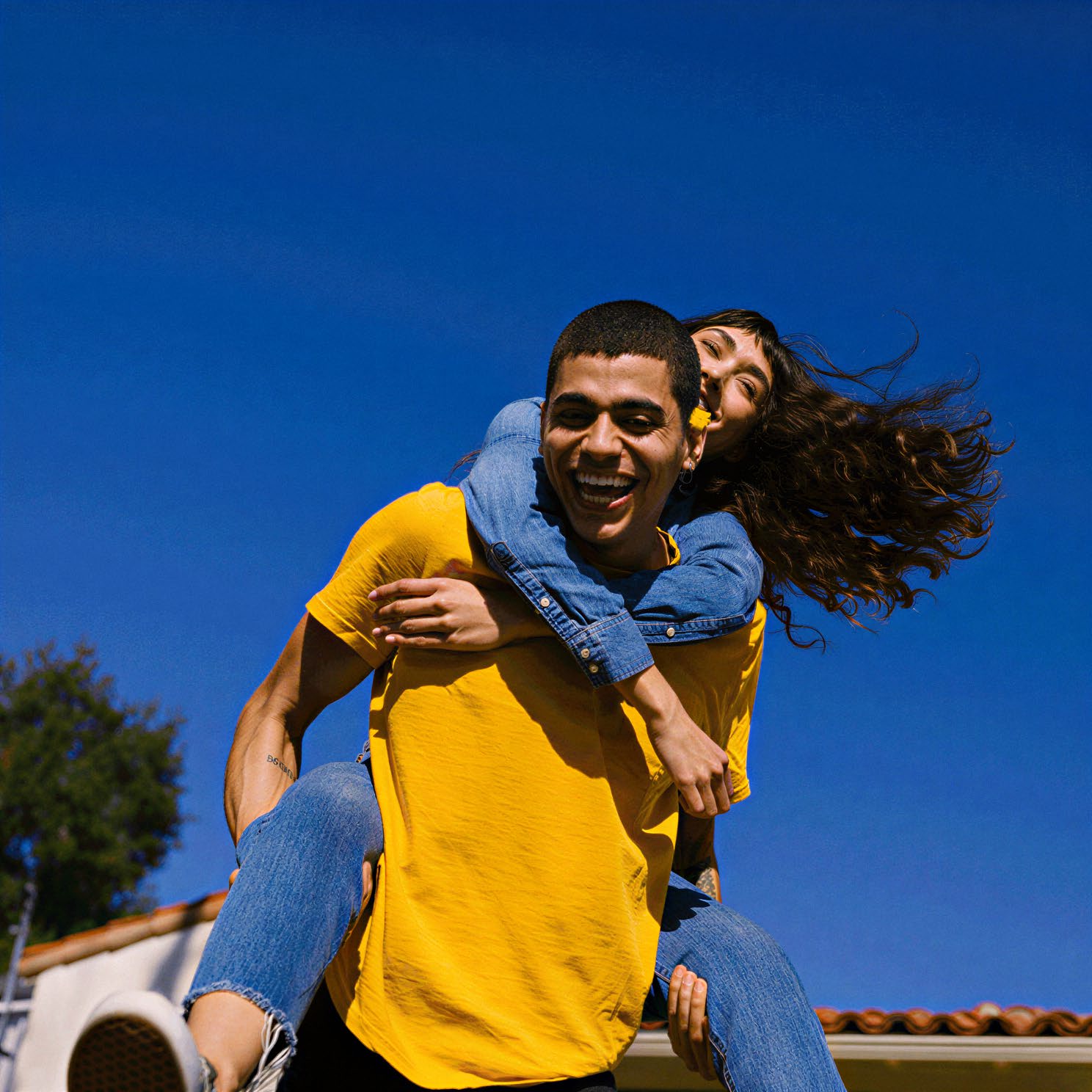 Casal sorrindo, um nas costas do outro, com céu azul ao fundo